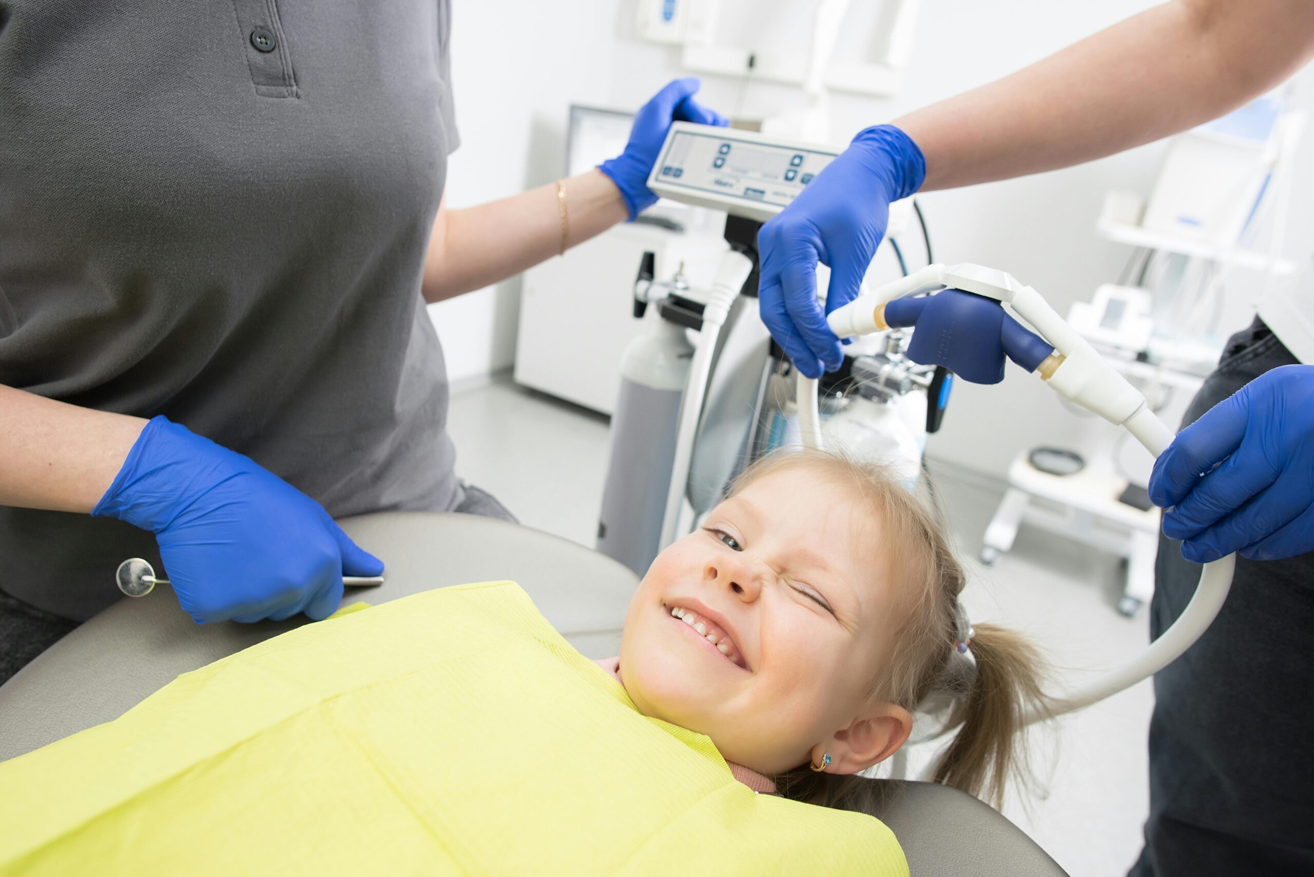 A cheerful child winks while sitting in a dental chair, surrounded by dental professionals.